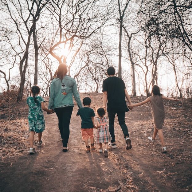 Eine Familie von sechs Personen geht Hand in Hand auf einem Weg durch einen kargen Wald.