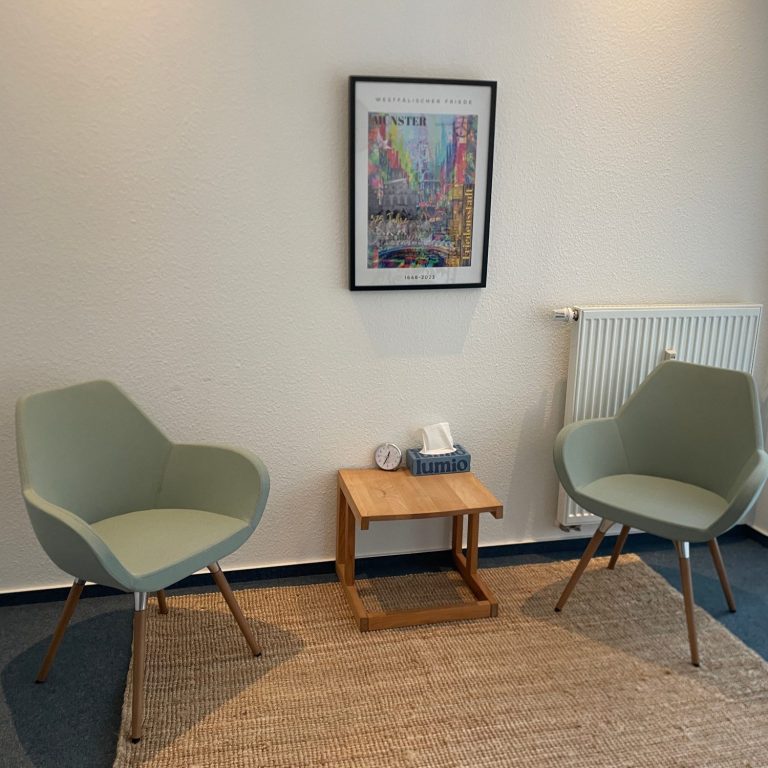 Beratungsraum Zwei Zwei grüne Stühle neben einem Tisch, mit einem Bild an der Wand und einem Heizkörper.